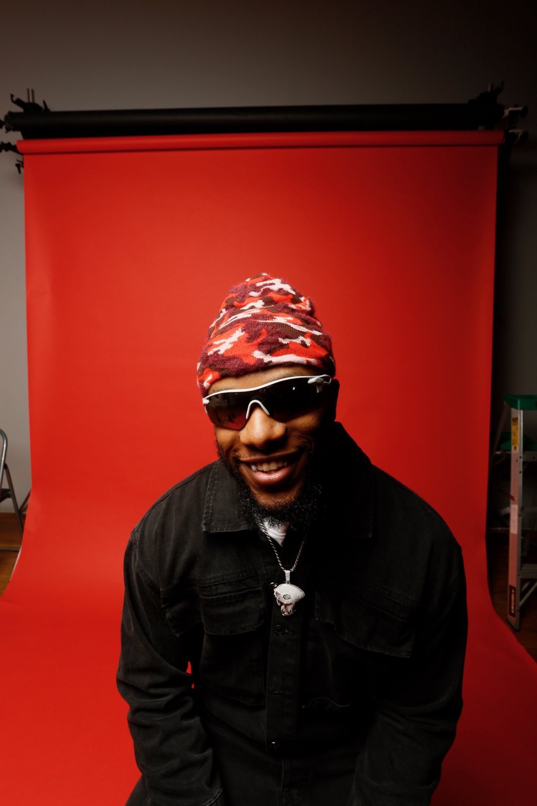 Smiling portrait in sunglasses and a camo beanie against a red backdrop